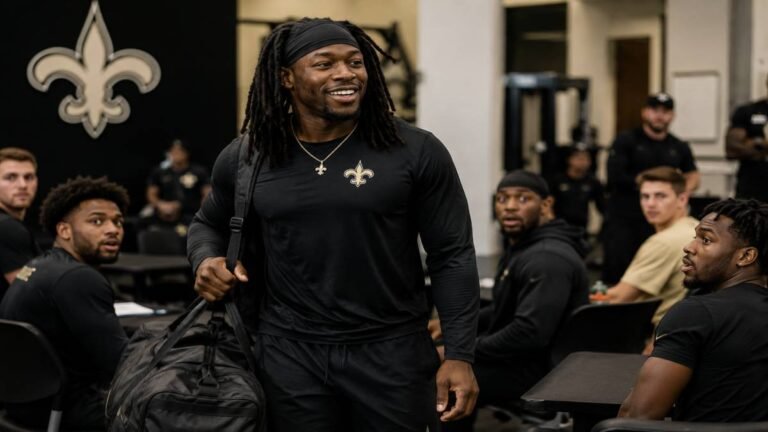Alvin Kamara, New Orleans Saints running back, walks into the Ochsner Sports Performance Center with a rolling bag and confident smile after a red-eye flight, as teammates react with surprise in the background during OTAs on June 3, 2026.