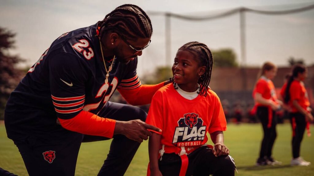 Devin Hester coaching young athletes during flag football drills on a Manchester school field as part of the Chicago Bears 2026 UK program. Kids actively engaged in pulls and routes on a sunny pitch with school buildings behind.
