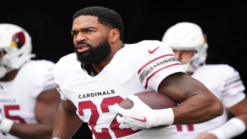 Arizona Cardinals quarterback Jacoby Brissett on the practice field during 2026 minicamp, serious expression with subtle contract debate and document overlays symbolizing his holdout negotiations.