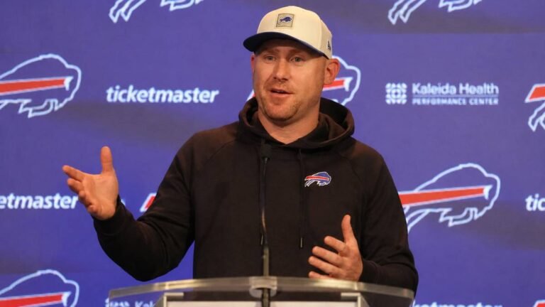 Buffalo Bills head coach Joe Brady sits thoughtfully with hand near his chin, wearing a team polo, against a backdrop featuring the Bills logo, conveying quiet determination during his first comments on Sean McDermott's firing.