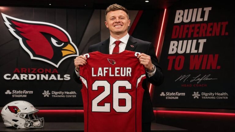 Arizona Cardinals head coach Mike LaFleur speaks with quarterbacks during 2026 minicamp practice, standing between two players in red jerseys and white helmets on a sunlit field in Arizona.
