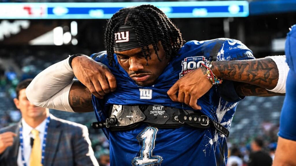 New York Giants wide receiver Malik Nabers leaps to make a spectacular catch in the end zone during a night game at MetLife Stadium, blue jersey shining under the lights with a roaring crowd behind him.