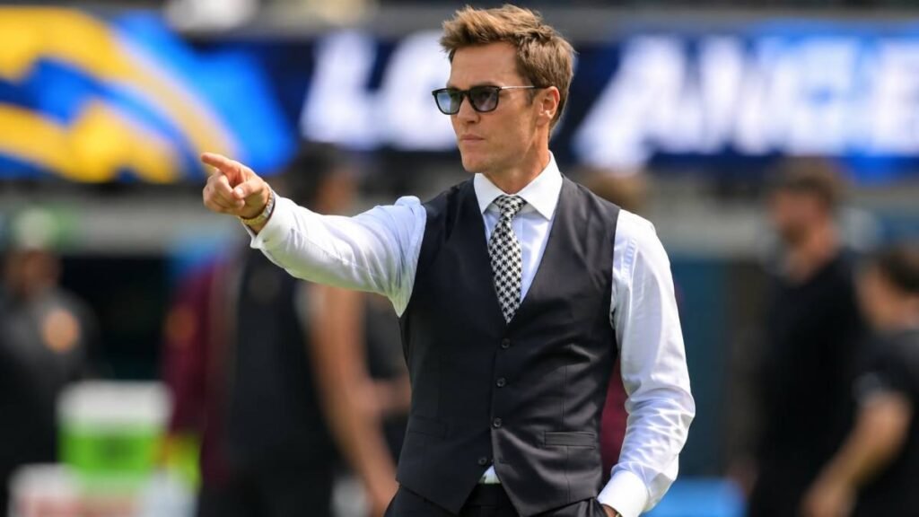Tom Brady stands on the field at Allegiant Stadium at sunset, wearing a Las Vegas Raiders jacket, symbolizing leadership and accountability as minority owner during the 2026 offseason.