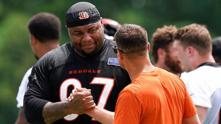 Cincinnati Bengals defensive players in orange and black gear drill on the Kettering Health Practice Fields with Paycor Stadium rising in the background on a clear summer day, capturing the fresh start for the unit after major 2026 offseason investments.