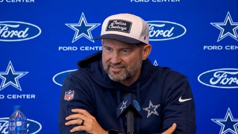 Dallas Cowboys head coach Brian Schottenheimer stands confidently outside AT&T Stadium wearing a navy team polo and cap with the Cowboys star logo, serious focused expression under bright Texas sunlight.