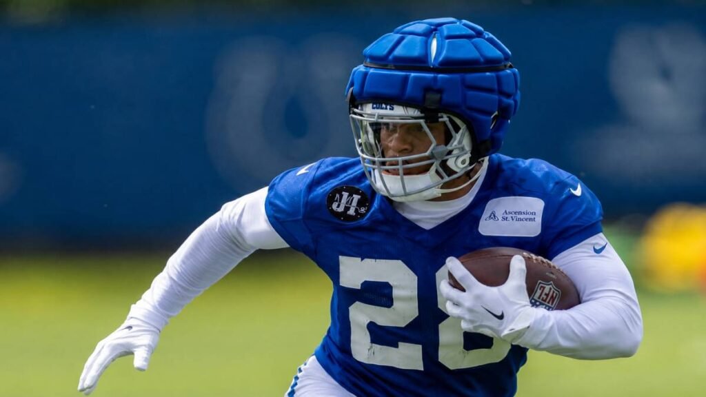 Jonathan Taylor #28 of the Indianapolis Colts sprints powerfully with the football during a 2026 NFL training camp practice under bright summer skies, with coaches and teammates visible in the background.