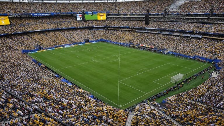 Wide view of the temporary natural grass pitch installed at MetLife Stadium for the 2026 FIFA World Cup final, with empty stands and bright sunlight highlighting the green surface.