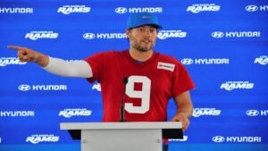 Los Angeles Rams quarterback Matthew Stafford in blue and gold uniform standing on SoFi Stadium field with floating golden dollar signs symbolizing NFL salary cap spending in 2026.