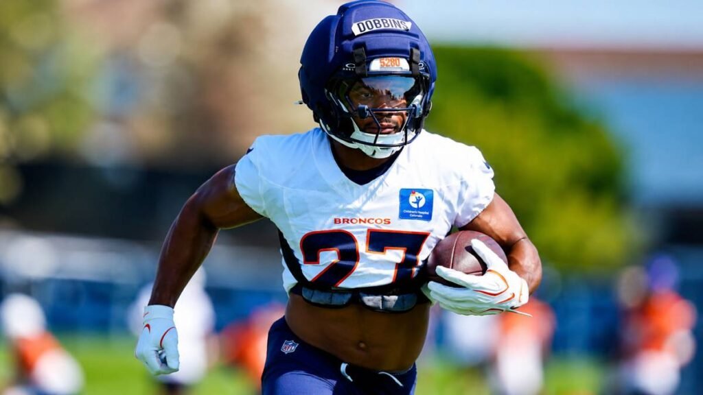 Denver Broncos running back J.K. Dobbins smiles and gestures passionately at a podium during his first training camp media session on July 31, 2026, wearing orange and navy No. 27 jersey with Rocky Mountains in the background.